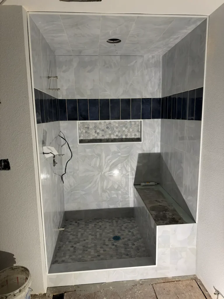 Walk-in shower under construction in Manitou Springs, CO, featuring light grey botanical wall tiles, a dark navy vertical accent band, a built-in bench, and hexagon mosaic floor tile by XCEL TILE.