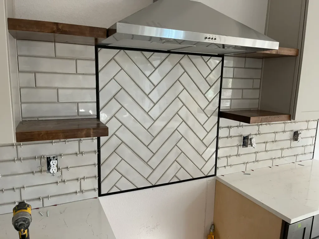 Kitchen remodel under construction showcasing a light grey herringbone tile accent wall perfectly framed beneath a stainless steel vent hood, flanked by standard subway tileTall, multi-tiered custom shower niche built into a light grey subway tile wall, featuring a light herringbone tile backer and white quartz shelves in Monument, CO, by XCEL TILE.