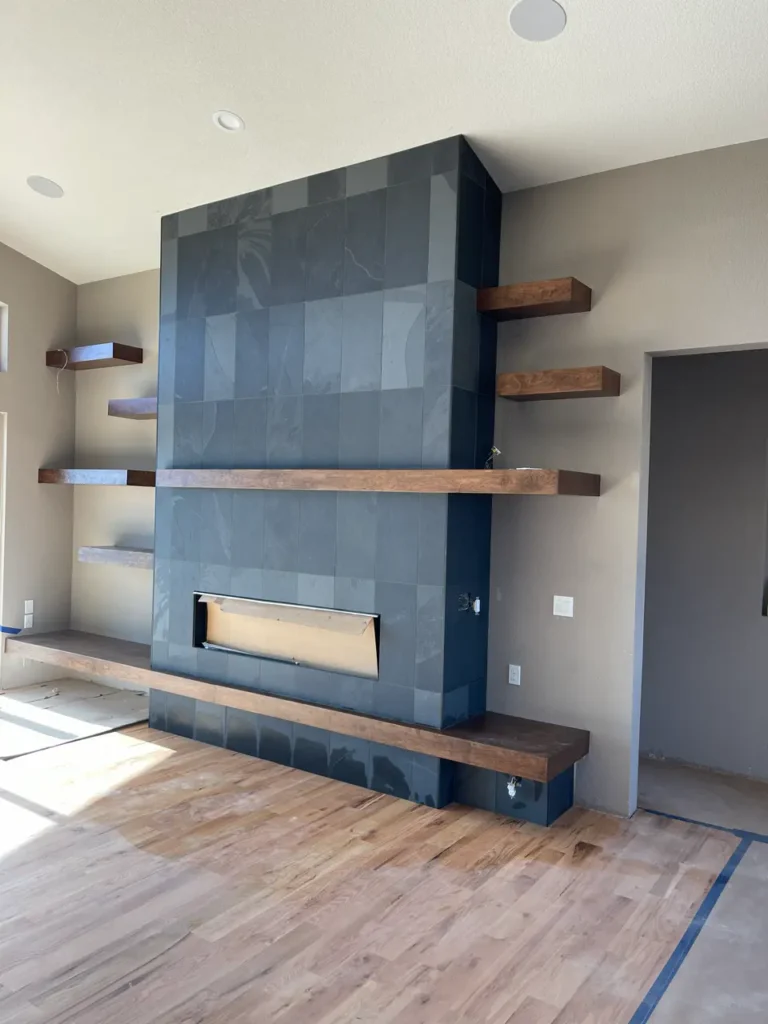 Wide view of a modern living room in Castle Rock, CO, featuring a massive custom fireplace wrapped in black slate tile, flanked by thick, natural wood floating shelves by XCEL TILE.