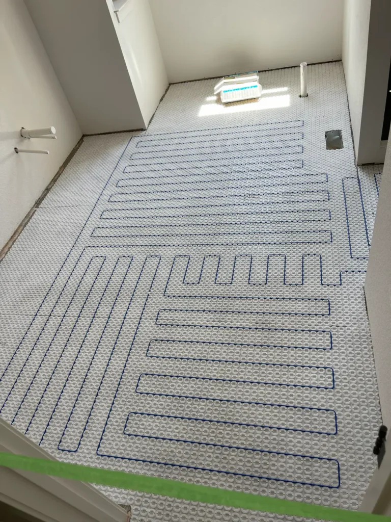 White uncoupling membrane with blue electric floor heating cables routed through it, laid out across a residential bathroom floor in Monument, CO, by XCEL TILE.