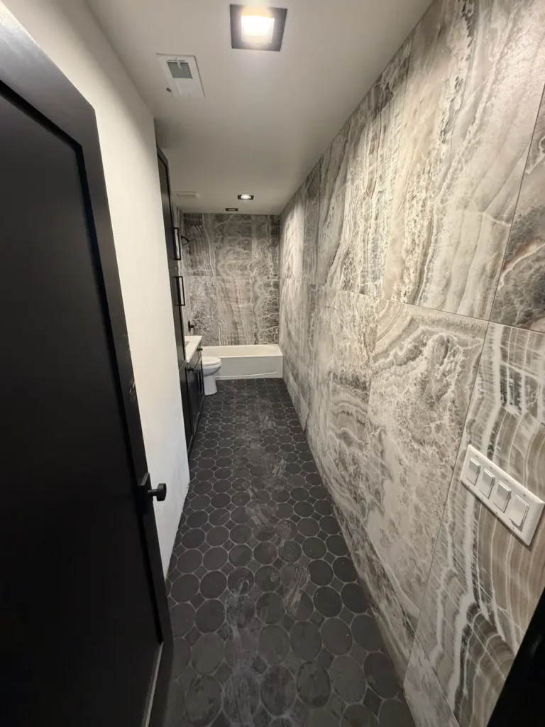 Narrow luxury bathroom space showcasing a continuous floor-to-ceiling installation of grey onyx-look porcelain wall tiles meeting a dark hexagon floor.