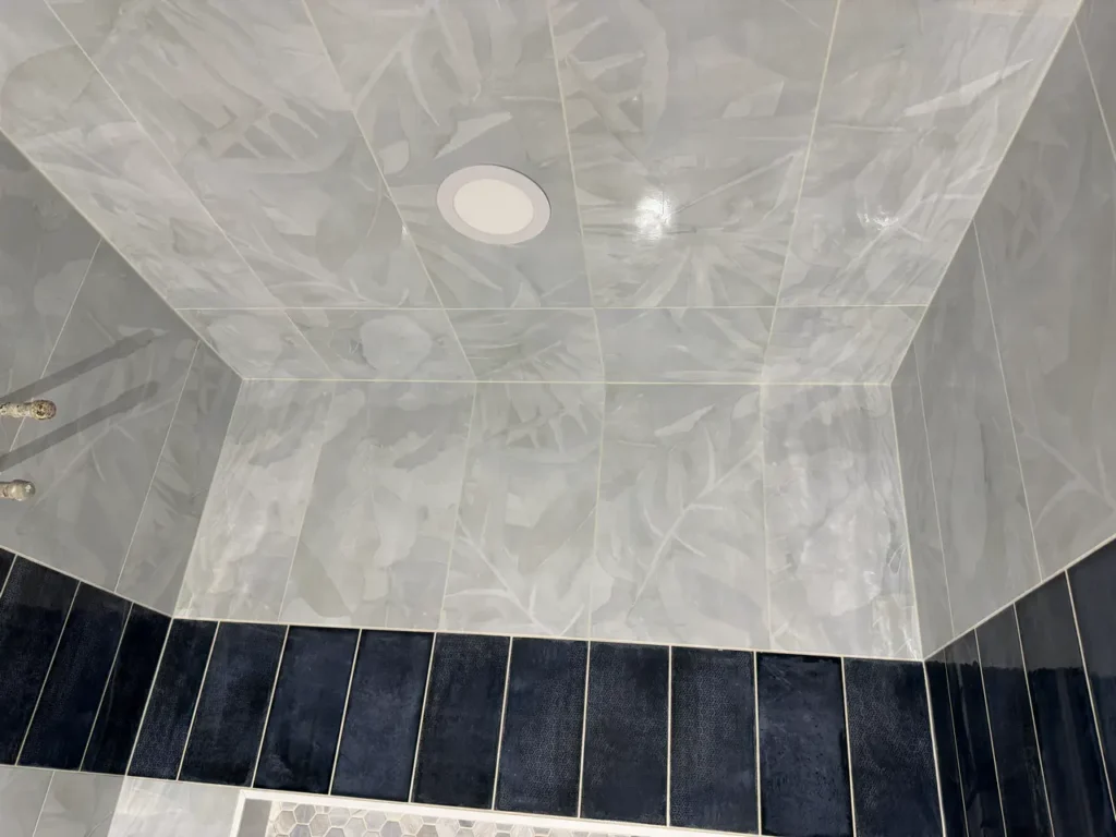 Looking up at a fully tiled shower ceiling in Manitou Springs, CO, featuring large-format light grey botanical pattern tiles, bordered by dark navy vertical accent tiles by XCEL TILE.