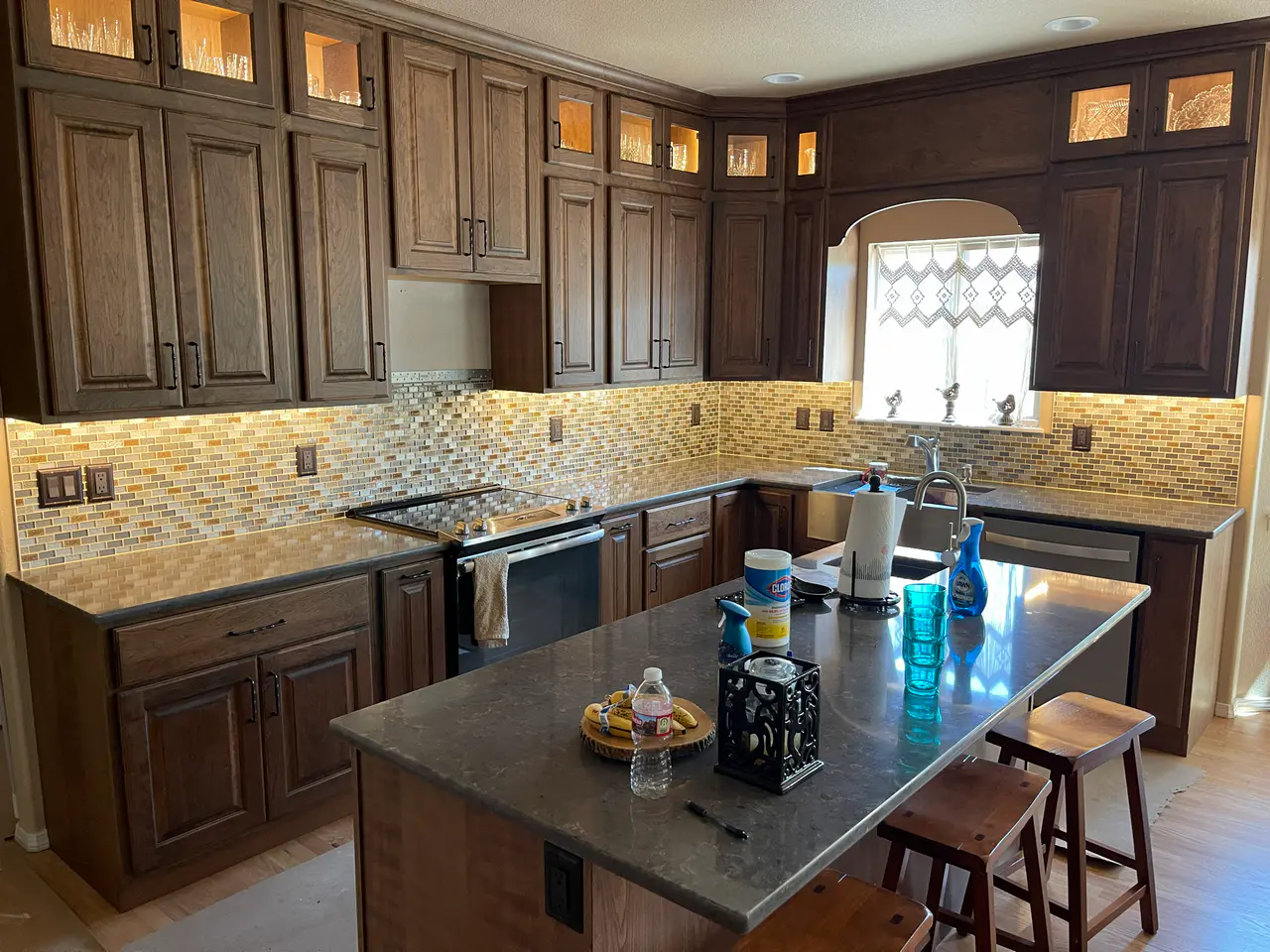 Wide view of a kitchen remodel Colorado Springs, CO, featuring dark wood cabinetry, a large center island, and a continuous multi-toned glass mosaic tile backsplash by XCEL TILE.