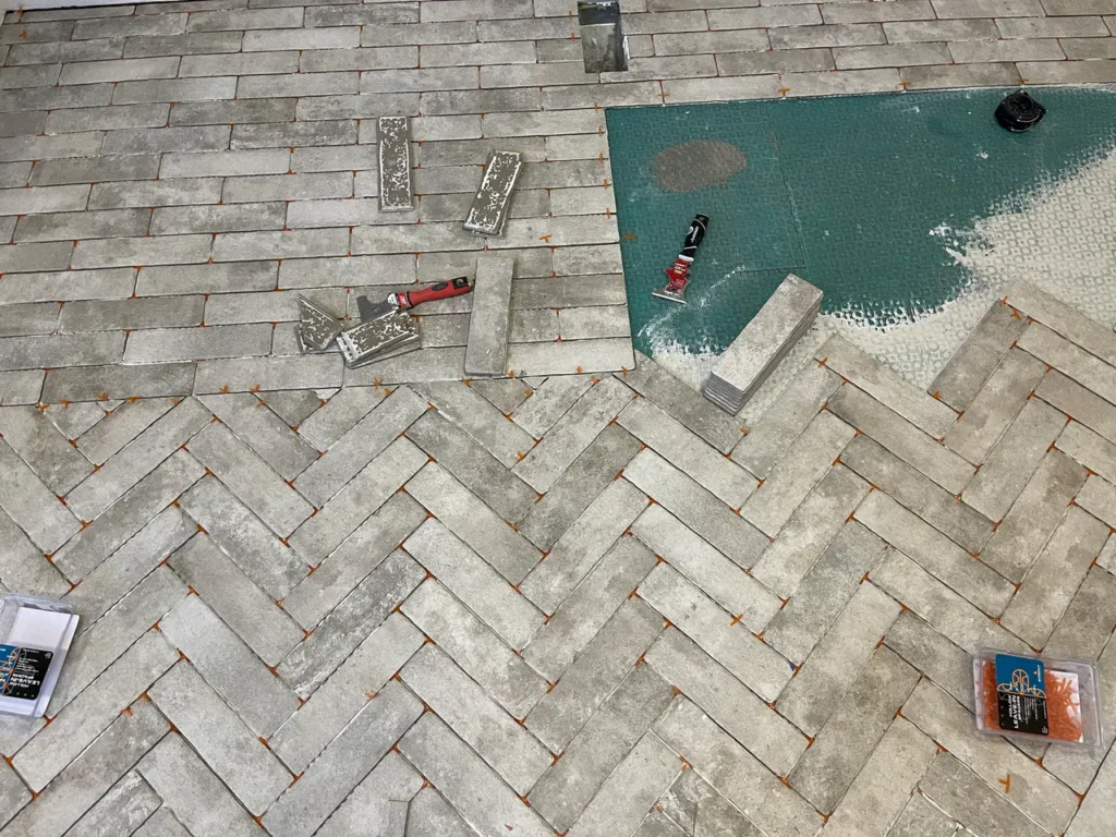 In-progress floor installation showing grey brick-look tiles being laid in a herringbone pattern over a green uncoupling membrane using orange spacers in Colorado Springs, CO, by XCEL TILE.