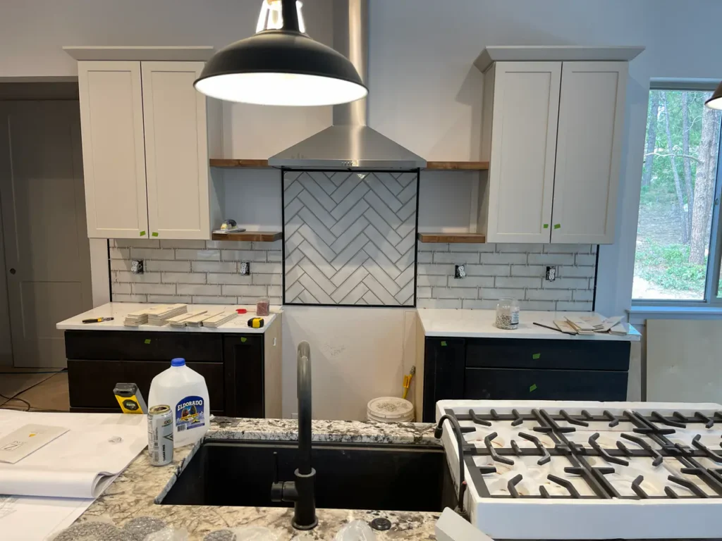 Kitchen remodel featuring a light grey herringbone tile accent wall framed beneath a stainless steel vent hood, centered between custom cabinetry and floating shelvesTall, multi-tiered custom shower niche built into a light grey subway tile wall, featuring a light herringbone tile backer and white quartz shelves in Monument, CO, by XCEL TILE.