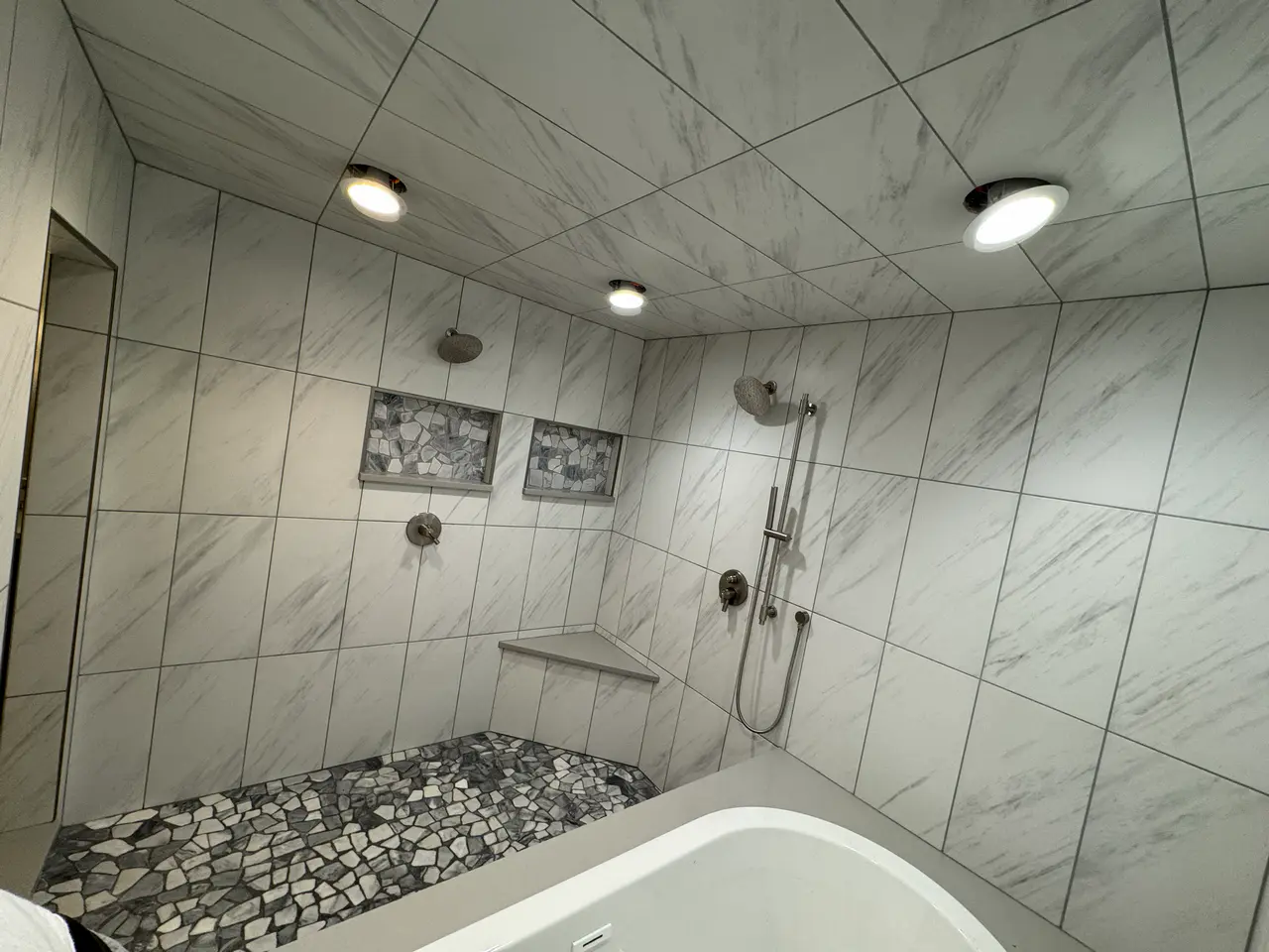 High-end wet room in the Broadmoor, CO, featuring white marble-look porcelain wall tiles, recessed niches, a corner bench, and a natural stone pebble floor by XCEL TILE.