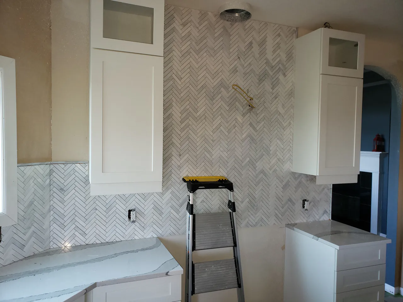 Kitchen wall under construction showcasing a floor-to-ceiling white marble herringbone tile installation centered between white upper cabinets over a quartz countertop. Custom Kitchen Backsplash Colorado Springs | XCEL TILE