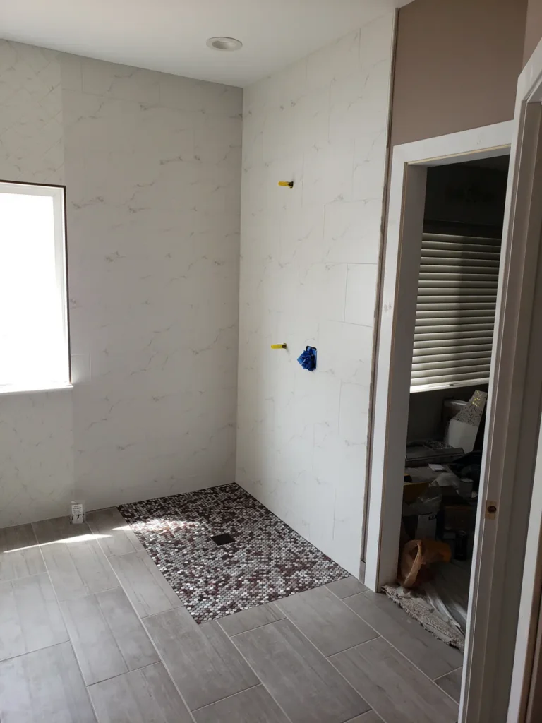 Bathroom remodel in progress in Parker, CO, showing large-format white marble-look porcelain tile installed on the walls, meeting a light wood-look tile floor and penny mosaic accent by XCEL TILE.