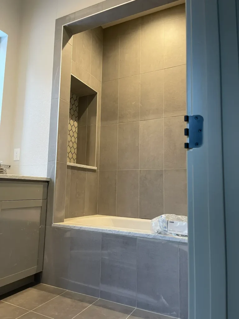 Framed view looking into a bathroom showing a bathtub surround clad in large-format, matte grey tile, featuring a tall, recessed storage niche with a decorative mosaic backer in Parker, CO, by XCEL TILE.