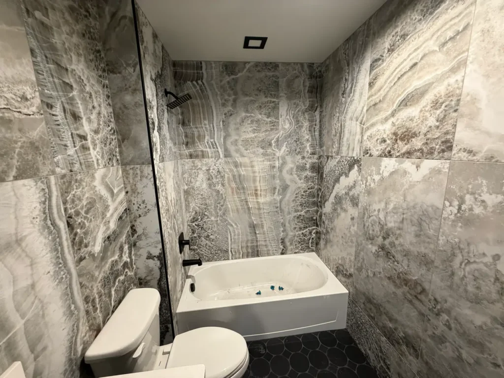 Bathroom featuring a bathtub and vanity surrounded by large-format grey onyx-look porcelain tiles on the walls and dark hexagon floor tiles.