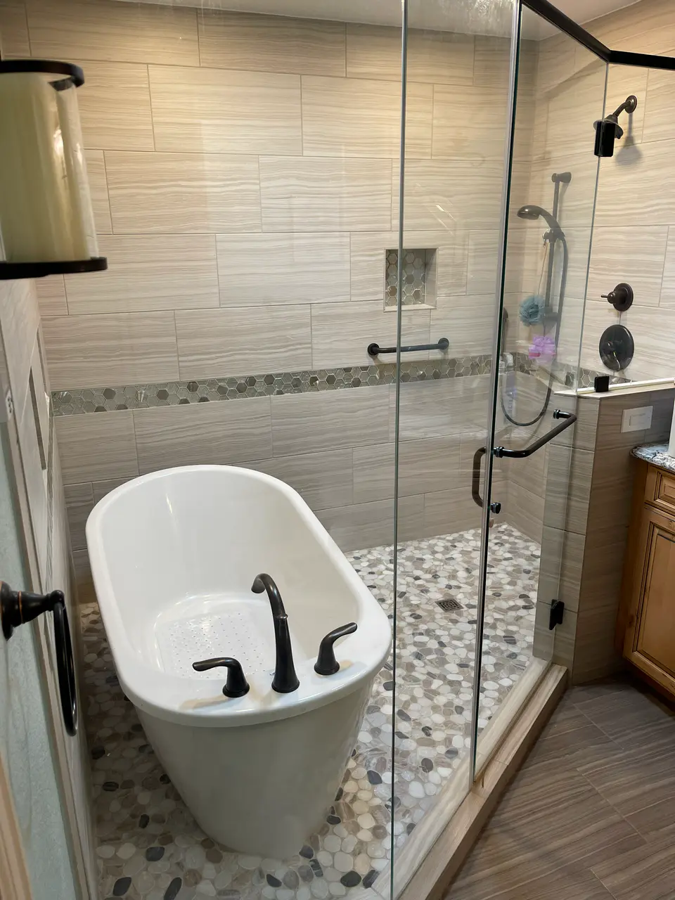 Luxury wet room featuring a freestanding soaking tub next to a glass-enclosed walk-in shower with neutral wood-look tile and stone pebble floor.
