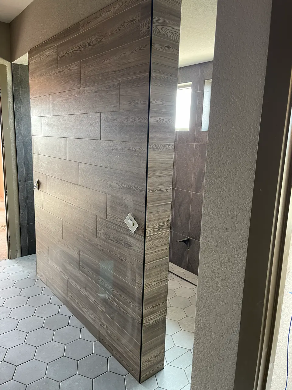 Custom bathroom partition wall completely wrapped in horizontally stacked wood-look tile, contrasting with a light grey hexagon floor tile.