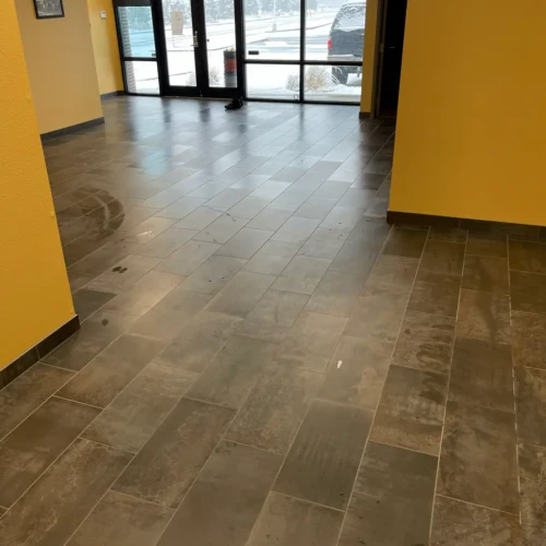 Wide view of a commercial office lobby in Colorado Springs, CO, featuring newly installed grey rectangular floor tile leading toward exterior glass entry doors by XCEL TILE.