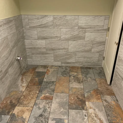 Grey rectangular wall tile installed as protective wainscoting above a multi-colored natural slate floor in a commercial restroom in Colorado Springs, CO, by XCEL TILE.