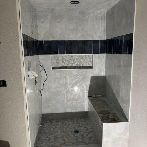 Walk-in shower under construction in Manitou Springs, CO, featuring light grey botanical wall tiles, a dark navy vertical accent band, a built-in bench, and hexagon mosaic floor tile by XCEL TILE.