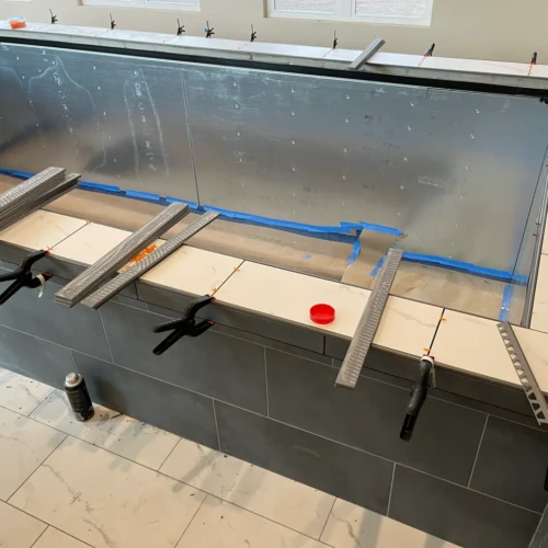 White marble-look pool coping tiles being installed and secured with heavy-duty clamps and leveling clips over a dark grey tiled pool wall in Parker, CO, by XCEL TILE.