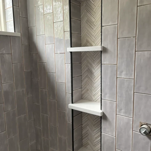 Tall, multi-tiered custom shower niche built into a light grey subway tile wall, featuring a light herringbone tile backer and white quartz shelves in Monument, CO, by XCEL TILE.