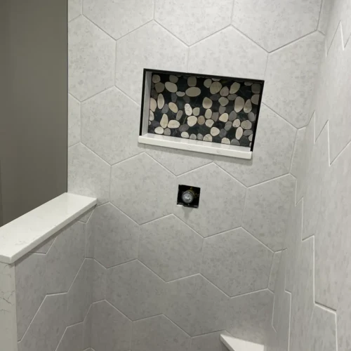 Close-up of a walk-in shower in Woodland Park, CO, featuring large light grey hexagon wall tiles and a recessed niche with pebble mosaic backing by XCEL TILE.