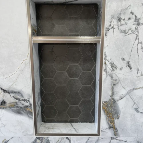 Close-up of a custom two-tiered shower niche in Monument, CO, featuring dark hexagon mosaic backing, framed by a bright brass metallic edge trim within marble-look wall tile by XCEL TILE.