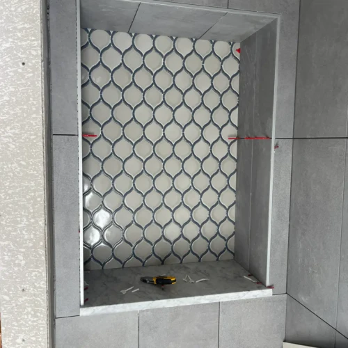 Close-up of a large custom shower niche built into a dark grey tile wall, featuring a delicate blue and white scallop mosaic backer and a white marble sill in Parker, CO, by XCEL TILE.