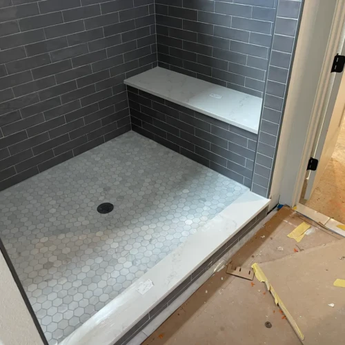 Walk-in shower under construction in Larkspur, CO, featuring a white hexagon marble mosaic floor, dark grey subway tile walls, and a built-in white quartz bench by XCEL TILE.