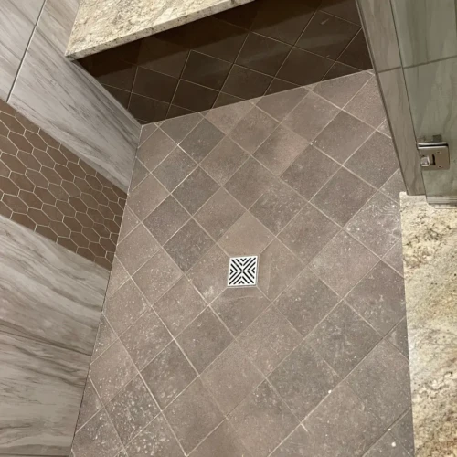 View looking down into a custom walk-in shower featuring a diagonally set stone-look floor tile, a square center drain, and a built-in bench with a quartz top and diamond-pattern tile face in Parker, CO, by XCEL TILE.