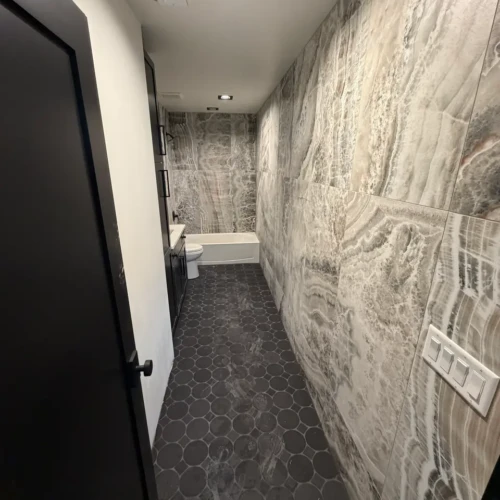 Narrow luxury bathroom space in Colorado Springs, CO, showcasing a continuous floor-to-ceiling installation of grey onyx-look porcelain wall tiles meeting a dark hexagon floor by XCEL TILE.