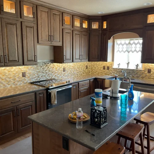 Wide view of a kitchen remodel Colorado Springs, CO, featuring dark wood cabinetry, a large center island, and a continuous multi-toned glass mosaic tile backsplash by XCEL TILE.