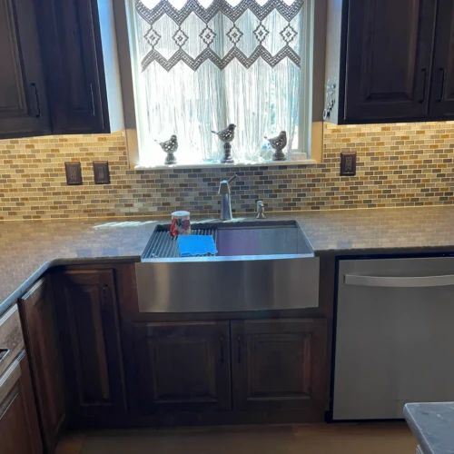 Kitchen sink area Colorado Springs, CO, featuring a stainless steel apron sink, dark wood cabinets, and a glass mosaic tile backsplash seamlessly wrapping around a sunlit window by XCEL TILE.