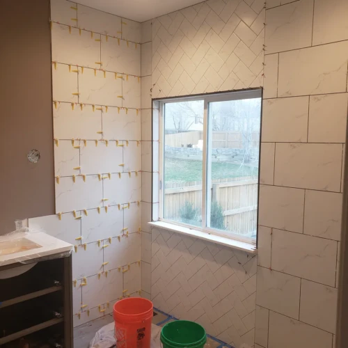 Bathroom walls under construction in Parker, CO, featuring white marble-look tiles installed in both a standard offset pattern and a complex herringbone layout around a window, held by yellow leveling clips by XCEL TILE.