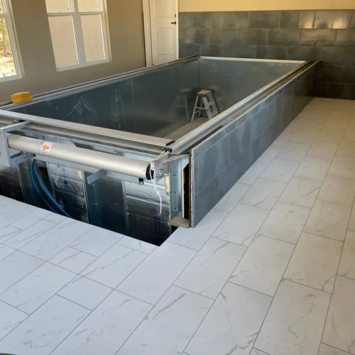 Wide view of a residential indoor pool area featuring a metal pool shell, dark grey tile surround, and bright white marble-look tile floors in Parker, CO, by XCEL TILE.