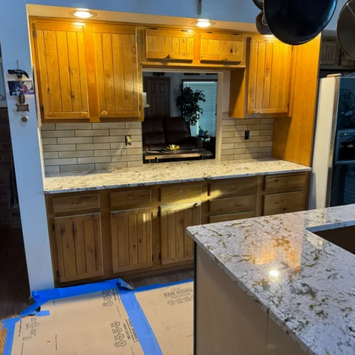Warm-toned residential kitchen remodel in Monument, CO, featuring light beige subway tile backsplash installed beneath natural wood cabinets, adjacent to a large granite-topped island by XCEL TILE.