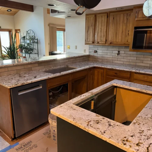 Wide view of a kitchen under construction, showing a light beige subway tile backsplash installed under natural wood cabinets and a two-tiered granite peninsula in Monument, CO, by XCEL TILE.
