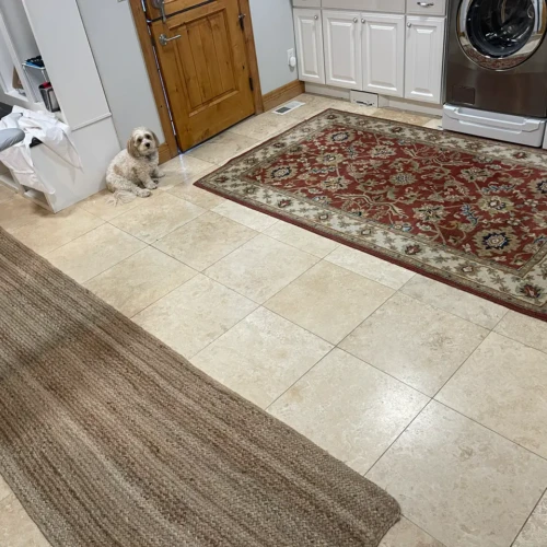 A "before" photo of a large residential laundry room featuring outdated beige square floor tiles prior to a custom remodel in Colorado Springs, CO, by XCEL TILE.
