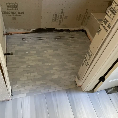 Walk-in shower pan under construction, featuring light grey subway tile flooring sloped toward a sleek linear drain against the back wall, surrounded by waterproof backing board in Parker, CO, by XCEL TILE.