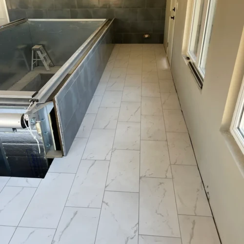 Seamless white marble-look tile flooring installed perfectly alongside an elevated indoor pool clad in dark grey rectangular tile in Parker, CO, by XCEL TILE.