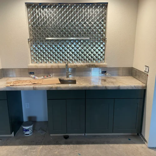 Custom residential bar area featuring dark green base cabinets, a light stone countertop, and a highly textured, reflective geometric tile accent wall surrounding a window in Parker, CO, by XCEL TILE.