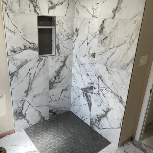 Walk-in shower enclosure in Monument, CO, finished in large-format white marble-look wall tiles, contrasting dark hexagon floor tiles, and a custom built-in niche by XCEL TILE.