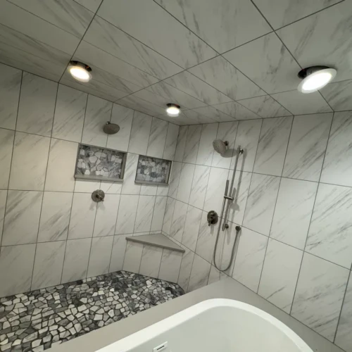High-end wet room in the Broadmoor, CO, featuring white marble-look porcelain wall tiles, recessed niches, a corner bench, and a natural stone pebble floor by XCEL TILE.