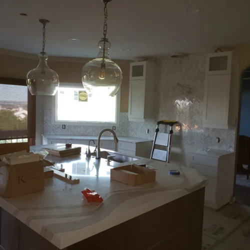 Wide view of a bright kitchen remodel in Colorado Springs, CO, featuring white cabinetry, a large island, and a continuous white marble herringbone tile backsplash by XCEL TILE.