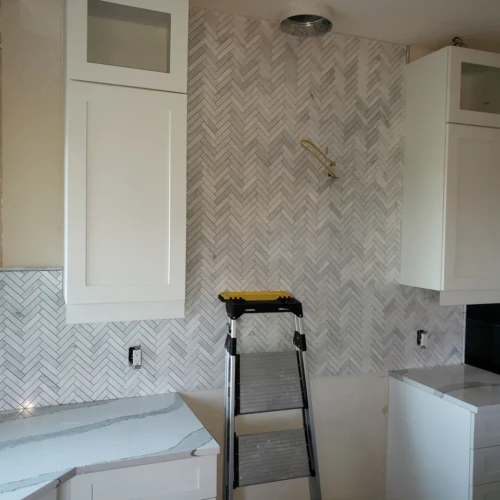 Kitchen wall under construction Colorado Springs, CO, showcasing a floor-to-ceiling white marble herringbone tile installation centered between white upper cabinets over a quartz countertop by XCEL TILE.