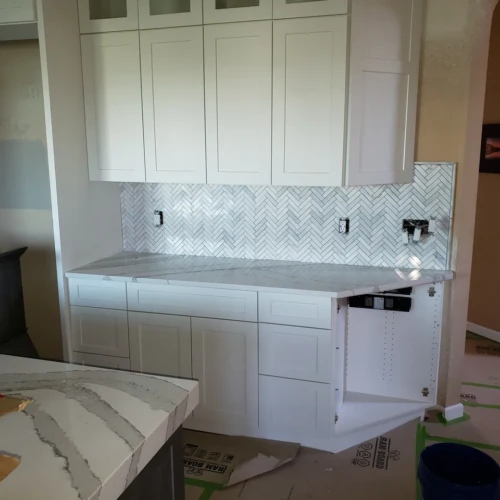 Completed section of a kitchen remodel Colorado Springs, CO, featuring white base and upper cabinets separated by a meticulously installed white marble herringbone tile backsplash by XCEL TILE.