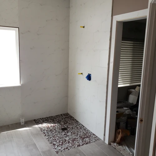 Bathroom remodel in progress in Parker, CO, showing large-format white marble-look porcelain tile installed on the walls, meeting a light wood-look tile floor and penny mosaic accent by XCEL TILE.
