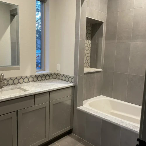 Modern residential bathroom featuring grey vanity cabinets, a white marble countertop, and a bathtub surround framed in large-format grey tile with a matching decorative mosaic backsplash in Parker, CO, by XCEL TILE.