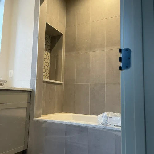 Framed view looking into a bathroom showing a bathtub surround clad in large-format, matte grey tile, featuring a tall, recessed storage niche with a decorative mosaic backer in Parker, CO, by XCEL TILE.