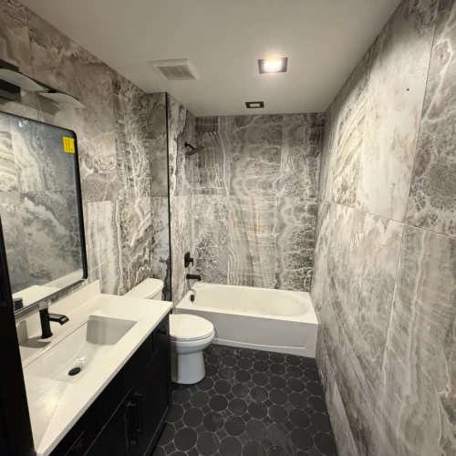 Bathroom in Colorado Springs, CO, featuring a bathtub and vanity surrounded by large-format grey onyx-look porcelain tiles on the walls and dark hexagon floor tiles by XCEL TILE.