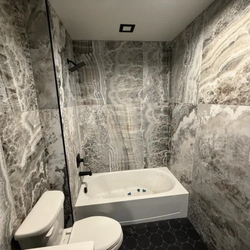 Bathroom in Colorado Springs, CO, featuring a bathtub and vanity surrounded by large-format grey onyx-look porcelain tiles on the walls and dark hexagon floor tiles by XCEL TILE.