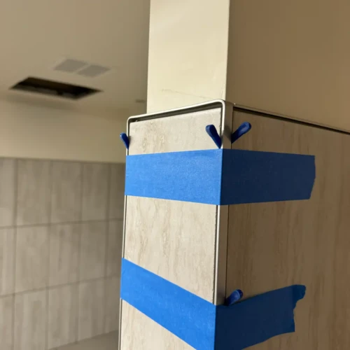 Close-up of light grey wall tile installation in a commercial restroom featuring a sleek metallic edge trim held in place with blue painter's tape in Colorado Springs, CO, by XCEL TILE.