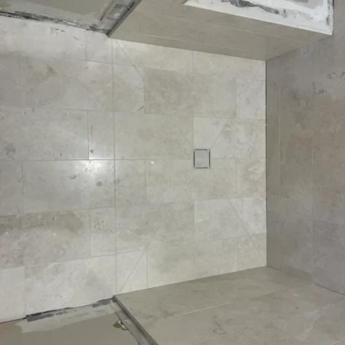 Top-down view of a custom shower pan in Colorado Springs, CO, featuring light stone-look tiles with precision envelope cuts sloping toward a square center drain by XCEL TILE.