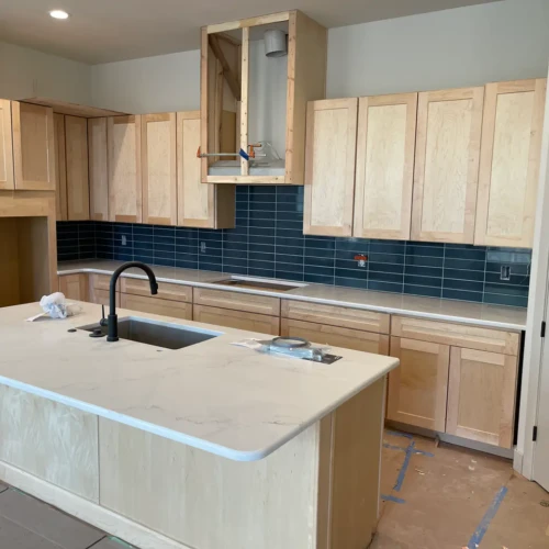 Wide view of a modern kitchen remodel in Parker, CO, featuring light wood cabinets, white quartz countertops, and a bold dark teal subway tile backsplash in a straight-stacked layout by XCEL TILE.